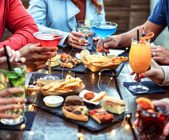 Group of friends enjoying appetizer drinking and eating in a bar - Close-up of hands of young people holding colorful cocktails in the happy hour time